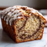 Close-up of a loaf of Cinnamon Roll Banana Bread with a thick white glaze drizzled on top and a visible cinnamon swirl inside.