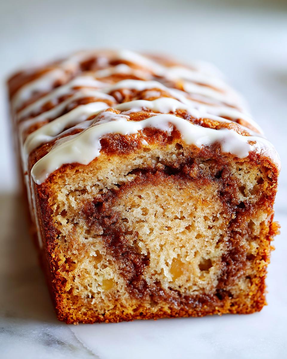 A close-up of a loaf of Cinnamon Roll Banana Bread, showing the moist crumb and cinnamon swirl, drizzled with white icing.