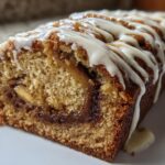 A close-up of a loaf of Cinnamon Roll Banana Bread, sliced to show the swirl and topped with white icing drizzle.
