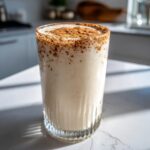 A tall glass filled with creamy Puerto Rican Coquito, topped generously with ground cinnamon.