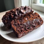 A close-up of a slice of rich, dark Double Chocolate Zucchini Bread next to the main loaf on a white plate.