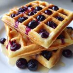 A close-up stack of fluffy Blueberry Waffles topped with bursting blueberries and drizzled with syrup.