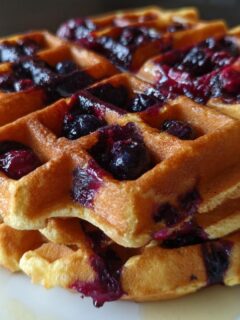 Close-up of a stack of fluffy Blueberry Waffles topped with warm blueberry compote and drizzled with syrup.