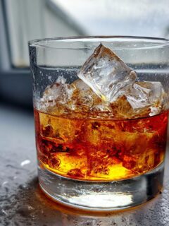 A close-up of a Godfather Cocktail, featuring amber liquor and large ice cubes, resting on a wet surface near a window.