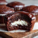 A close-up, bitten cross-section of a Homemade Hostess Cupcakes showing rich chocolate cake, white cream filling, and shiny chocolate glaze.
