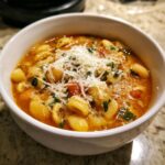 Close-up of a white bowl filled with rich Pasta Fagioli soup, topped generously with grated Parmesan cheese and fresh herbs.