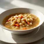 A close-up of a white bowl filled with hearty White Chickpea Chili featuring chickpeas, carrots, and onions in a savory broth.