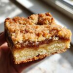 A close-up, handheld view of a thick, layered Apple Snickerdoodle Bars slice showing the crumbly topping and gooey apple filling.