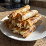 A stack of golden brown Baked Cream Cheese Chicken Taquitos with visible creamy filling on a white plate.