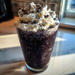 A tall glass filled with a dark, icy Black Halloween Milkshake, topped with whipped cream and chocolate sprinkles.