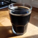 Close-up of a cold, dark Black Milkshake in a textured glass sitting on a sunlit wooden countertop.