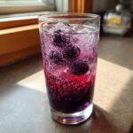 A tall glass filled with fizzy Blackberry Spider Soda, ice cubes, and whole blackberries floating in the purple liquid.
