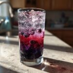 A tall glass filled with Blackberry Spider Soda, ice, and whole blackberries, showing vibrant purple and red layers.