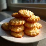 A stack of golden brown Buttery Spritz Cookies with ridged tops resting on a white plate near a window.