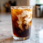 Close-up of a tall glass filled with Campfire Cola Dirty Soda, showing rich cream swirling dramatically into the dark soda over ice.