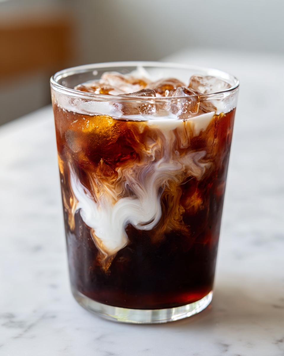 Close-up of a glass containing an iced, dark beverage with white cream swirling dramatically into the liquid, resembling a Campfire Cola Dirty Soda.