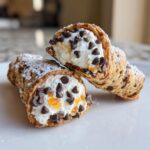 Two freshly made Cannoli Cookies filled with sweet cream, mini chocolate chips, and candied orange peel, dusted with powdered sugar.