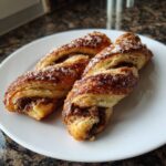 Two golden-brown Chocolate Chip Cookie Dough Twists sprinkled with coarse sugar on a white plate.