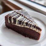 A perfect slice of Chocolate Spiderweb Cheesecake with dark chocolate ganache and white drizzle on a white plate.