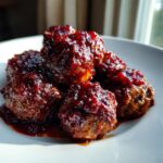Close-up of several glistening Christmas Cranberry Meatballs coated in a thick, dark red cranberry glaze on a white plate.