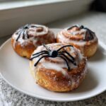 Three glazed Creepy Crawly Cinnamon Rolls decorated with black plastic spiders on a white plate.