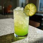 A tall glass filled with a bright green, fizzy Ectoplasm Elixir Mocktail, topped with crushed ice and a lime wheel.