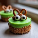 A close-up of one of the Frankenstein Mini Cheesecakes featuring a bright green top, graham cracker crust, and pretzel 'horns' with candy eyeballs.