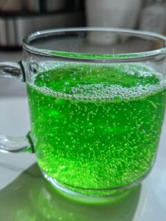 Close-up of fizzy, bright green Goblin Green Punch served in a clear glass mug on a white counter.
