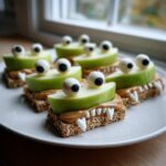 Several spooky Green Apple Monsters made with apple slices, peanut butter, candy eyes, and teeth on a white plate.