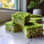 Three stacked squares of bright green Frankenstein Rice Krispie Bars sitting on a white marble surface.