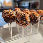 A close-up of several chocolate-dipped Halloween Cake Pops covered in orange, black, and white sprinkles.