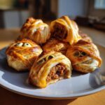 A pile of golden-brown Halloween Mummy Meatballs wrapped in pastry with olive eyes, one cut open showing the meat filling.