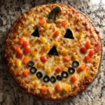 A fully baked Jack O Lantern Halloween Pizza decorated with colorful peppers and black olives on melted cheese.