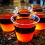 Close-up of layered orange and black Halloween Jello Shots in clear plastic cups on a granite surface.
