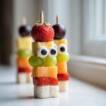 Close-up of a colorful Monster Fruit Skewer featuring a strawberry head, candy eyes, grapes, and melon cubes.