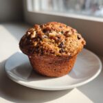 A large, freshly baked Monster Muffin topped with walnuts and chocolate chips, sitting on a small white plate.
