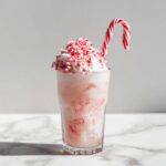 A tall glass filled with a pink Peppermint Christmas Float, topped with whipped cream and crushed candy cane, garnished with a whole candy cane.