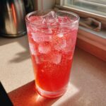 A tall glass filled with a bright red, fizzy Pink Potion Drink and ice cubes, glistening in the sunlight.