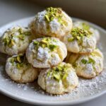 A stack of soft, round Pistachio Wedding Cookies dusted heavily with powdered sugar and topped with chopped green pistachios.