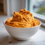 A white bowl filled high with fluffy, bright orange Pumpkin Cheesecake Dip, set on a marble counter near a window.
