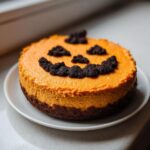 A vibrant orange Pumpkin Cheesecake Jack O Lantern sitting on a white plate, decorated with dark cookie crumbs for the face.