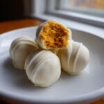 A stack of white chocolate coated Pumpkin Cheesecake Truffles, one bitten open revealing the orange filling.