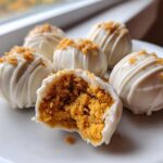 A white plate holds several Pumpkin Cheesecake Truffles coated in white chocolate, one is broken open showing the orange filling.