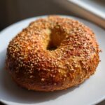 A golden-brown, sesame seed-crusted Monster Bagel resting on a white plate, highlighted by natural light.