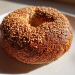 A close-up, sunlit photo of a perfectly baked Monster Bagel heavily coated in golden sesame seeds on a white plate.
