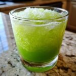 Close-up of a bright green, icy Slime Punch Mocktail filling a clear glass, resting on a granite countertop.