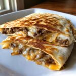 Close-up of two halves of golden-brown Smashburger Quesadillas overflowing with melted cheese, ground beef, and onions.