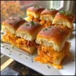 Close-up of several Spicy Buffalo Chicken Sliders overflowing with orange sauce and melted cheddar cheese on a white platter.