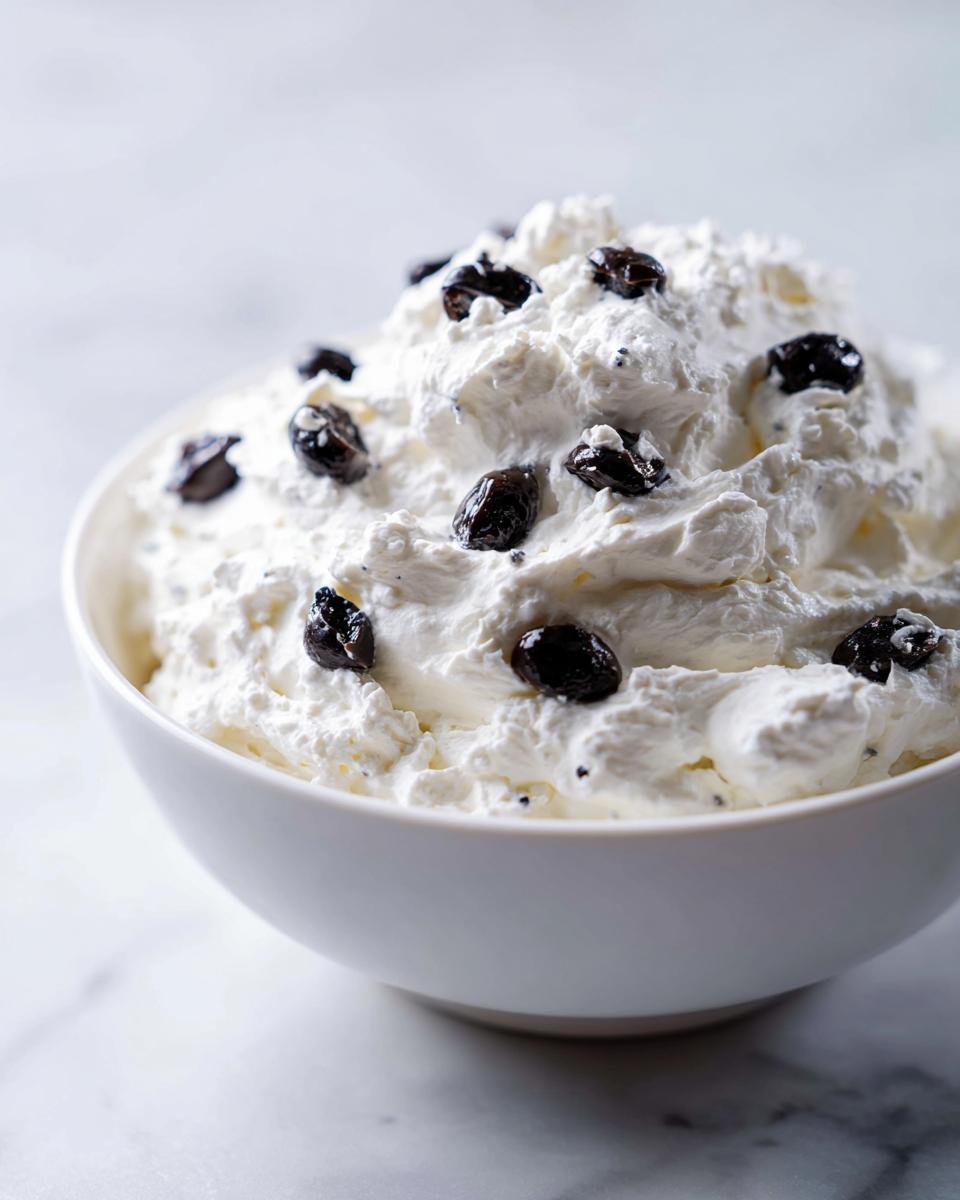 Close-up of a white bowl filled with fluffy Spider Egg Ranch Dip topped with black olive pieces.