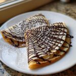 Two folded Spiderweb Crepes drizzled with dark chocolate sauce and dusted heavily with powdered sugar, served on a white plate.